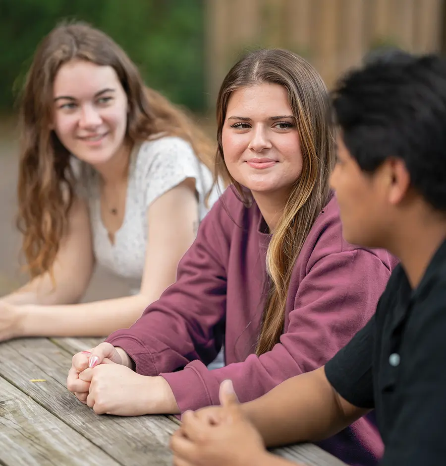 Portrait photograph close-up outdoor view of first generation students of CofC cofounders Katie Hughes (left) and Molly Moloney as they are both listening to another guy in a black polo top shirt speaking as all of them are at a wooden table