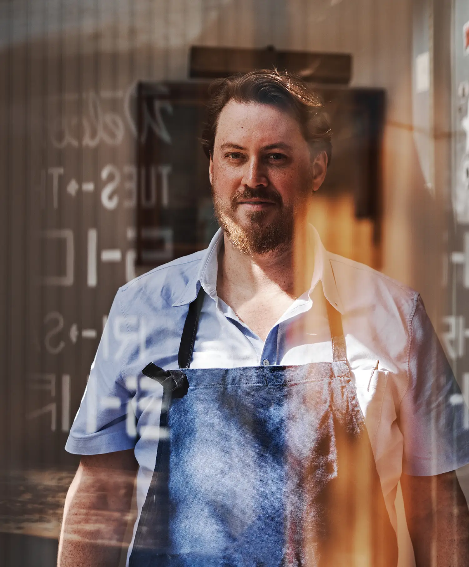 A man wearing a blue shirt and apron stands behind a window with text reflections.