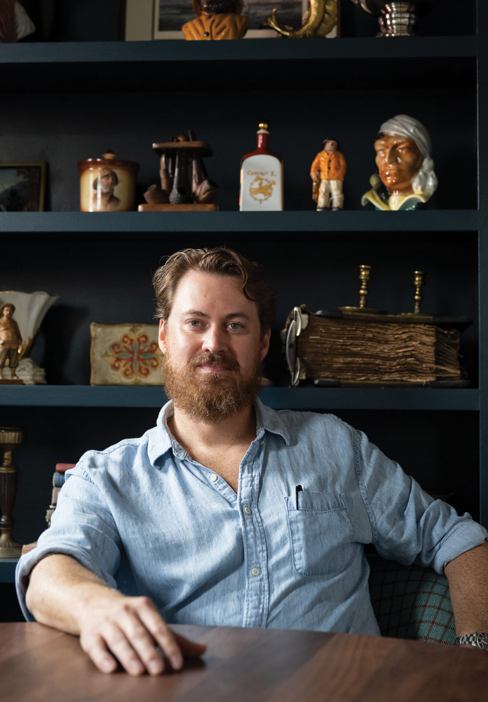 A bearded man in a light blue shirt sits at a wooden table with a dark bookshelf of eclectic items behind him.