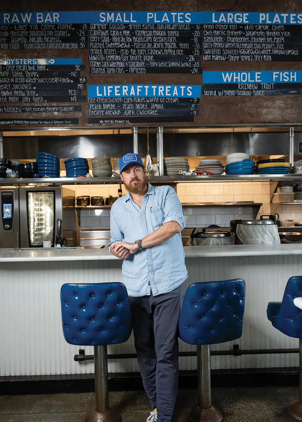 A man stands in front of a seafood bar counter with blue stools and chalkboard menus above.