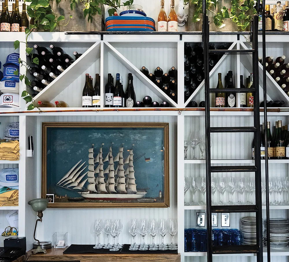 Shelving unit with wine bottles, a framed ship painting, and glassware, featuring a ladder and decorative vines.