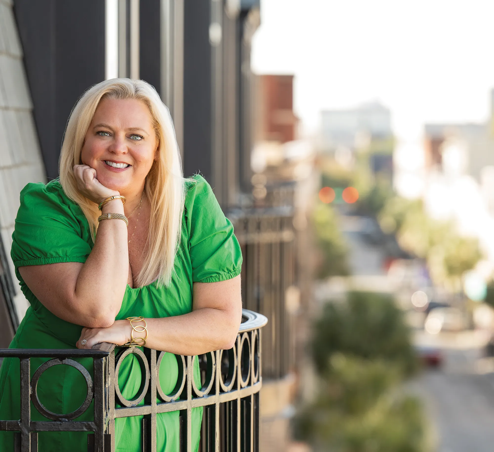 Person in a green dress leaning on a balcony railing with an urban background.