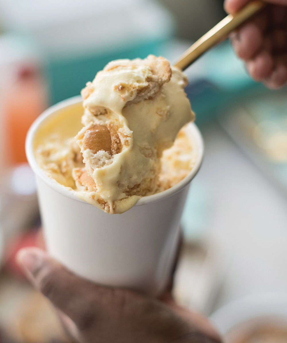 Close-up of ice cream with cookie chunks in a white cup, held by a hand with a gold-handled spoon.