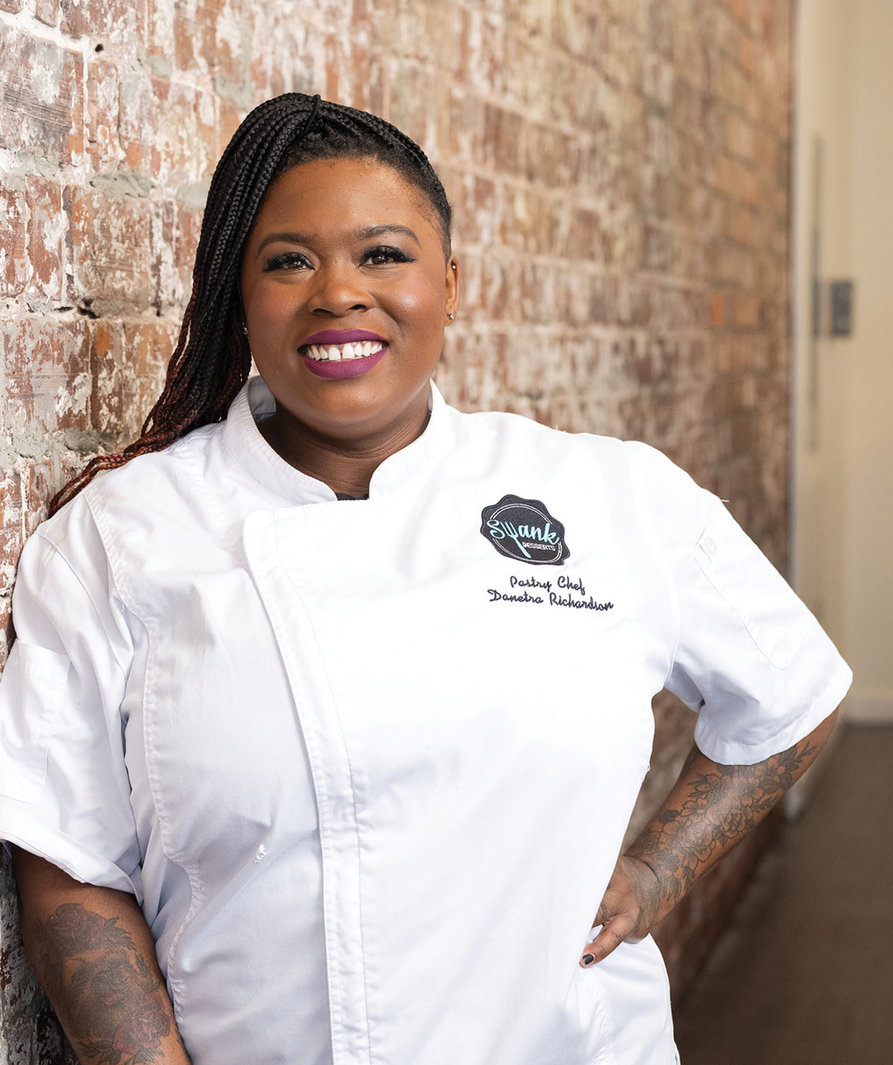 Person in a white chef's coat smiling against a brick wall.