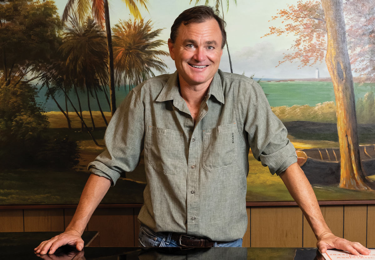 Man standing in front of a tropical mural, smiling with hands on a surface.