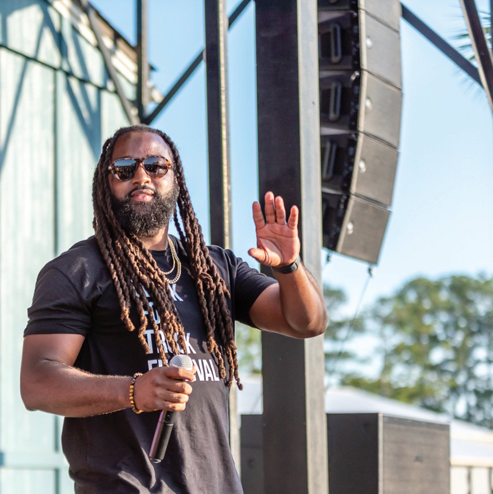 Man with long braids and sunglasses holding a microphone at an outdoor event.