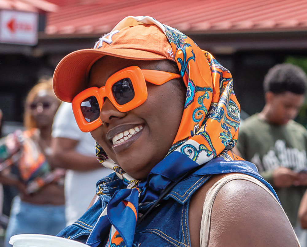 Person smiling with orange sunglasses, cap, and colorful headscarf.