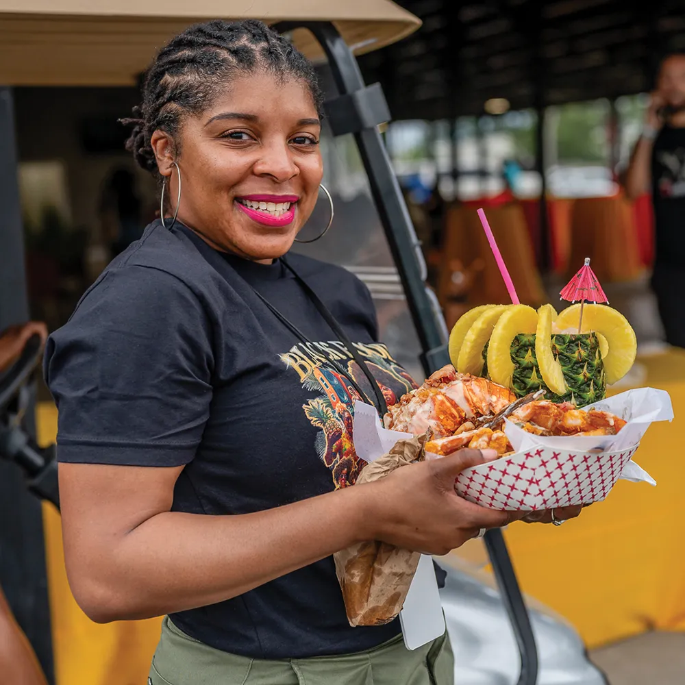 A woman with braided hair wearing a black T-shirt and hoop earrings, smiling while holding a tray with a pineapple cup and a dish topped with food, at an outdoor event.