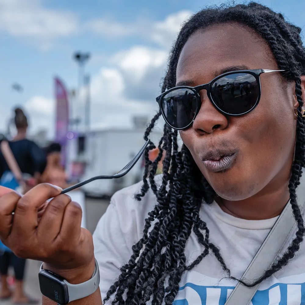 A woman wearing sunglasses and a white T-shirt, playfully eating with a fork at an outdoor event, with a blurred background of people and tents.