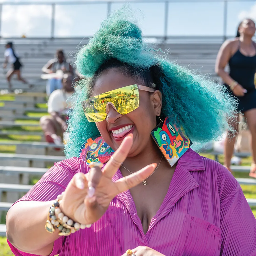 A woman with vibrant turquoise hair and oversized reflective sunglasses, smiling and holding up a peace sign while wearing colorful earrings and a purple outfit at an outdoor event.