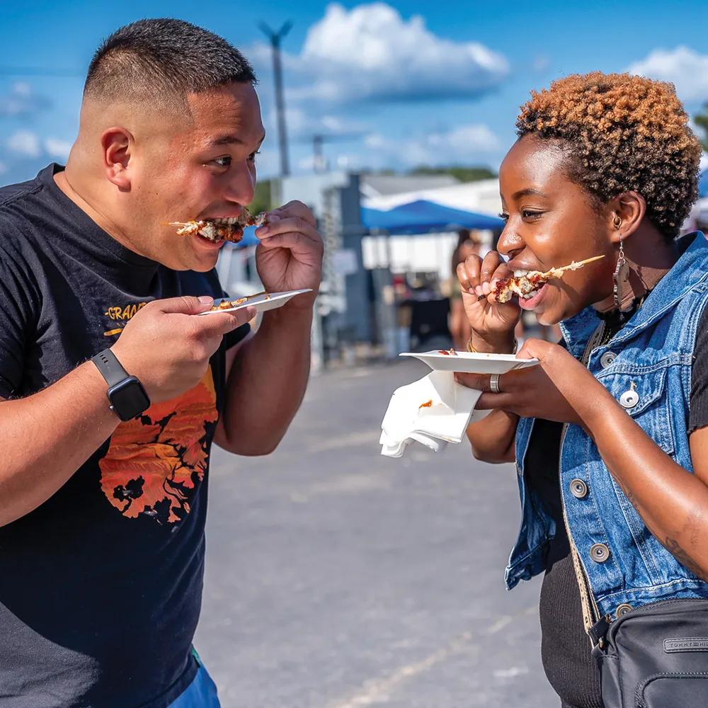 A man and woman enjoying food at an outdoor event, each holding plates and eating enthusiastically, with a bright and sunny atmosphere in the background.