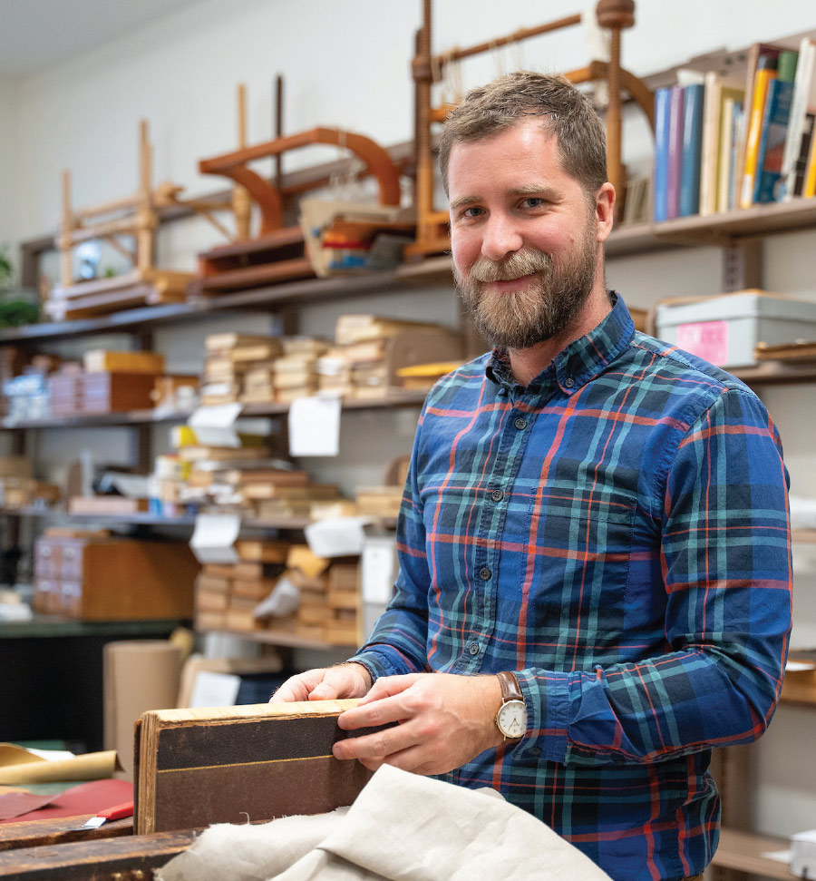 James Davis in blue, red, and green flannel at table with hands on book with full book shelves behind him