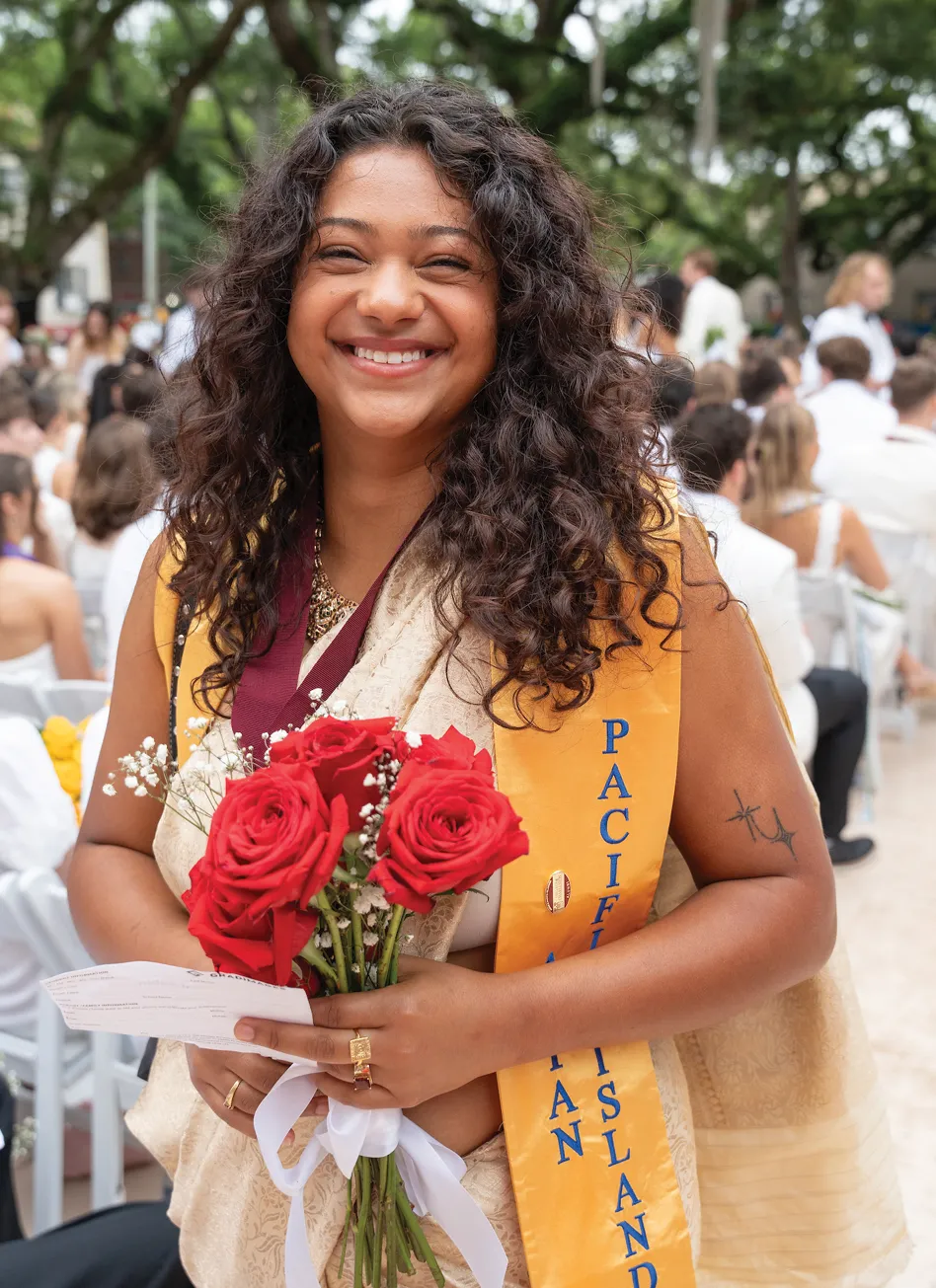 A smiling graduate with long curly hair wears a traditional cream sari and a yellow stole labeled “Pacific Islander.” She holds red roses and stands among a crowd of seated graduates outdoors.