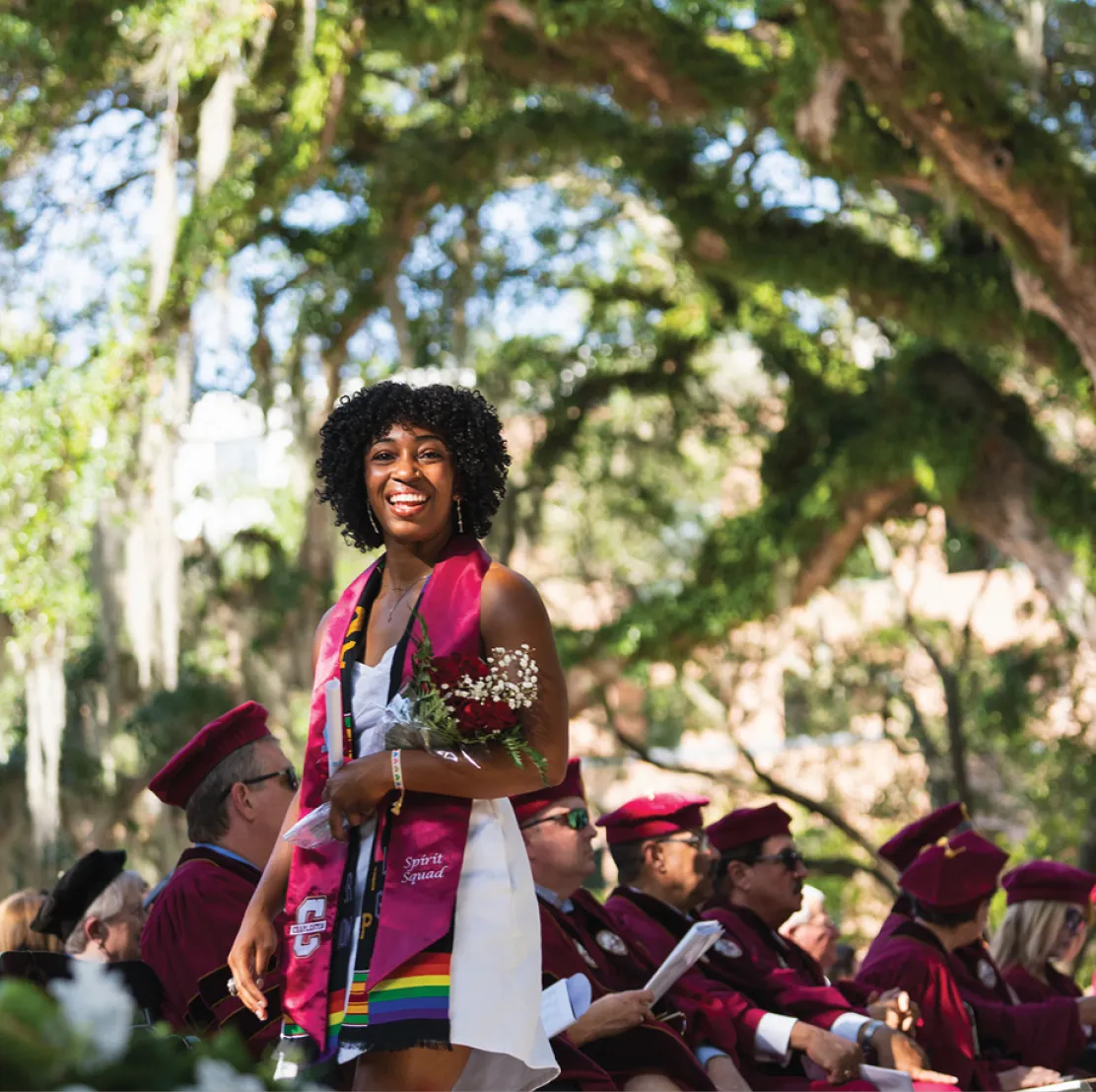 A smiling graduate wearing a maroon stole and a rainbow stole stands with a bouquet of red roses and white flowers. Faculty in maroon academic regalia are seated behind her under large oak trees.