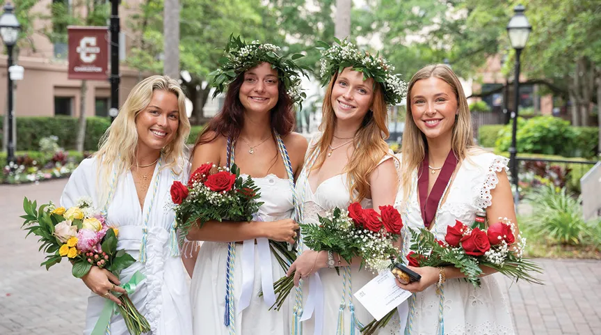 Four graduates in white dresses and leafy flower crowns hold colorful bouquets and smile at the camera while standing on a brick path lined with trees and campus buildings.