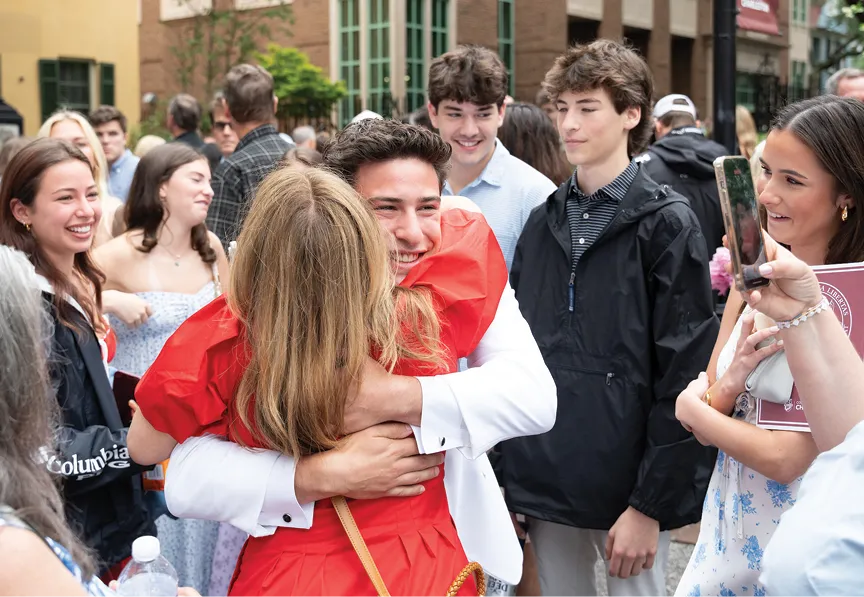 A graduate in a white tuxedo hugs a woman in a red dress while smiling broadly. They are surrounded by a happy group of family and friends capturing the moment with a smartphone.