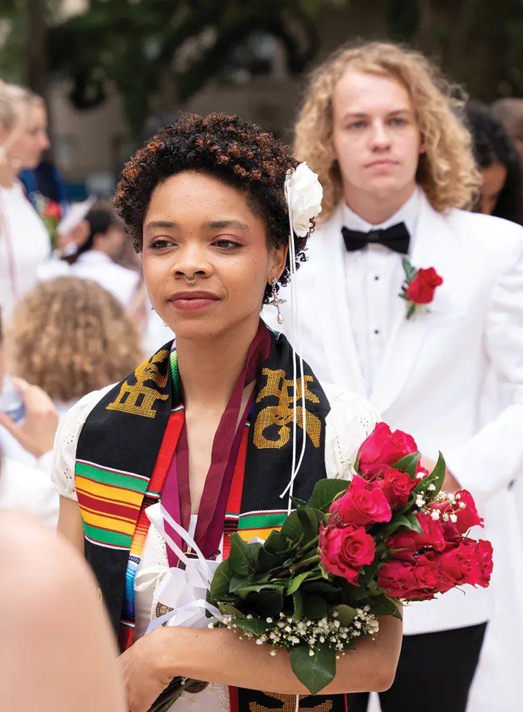 A graduate wearing a Kente cloth stole, maroon medal ribbon, and white lace dress stands holding a bouquet of red roses. Another graduate in a white tuxedo stands behind her.