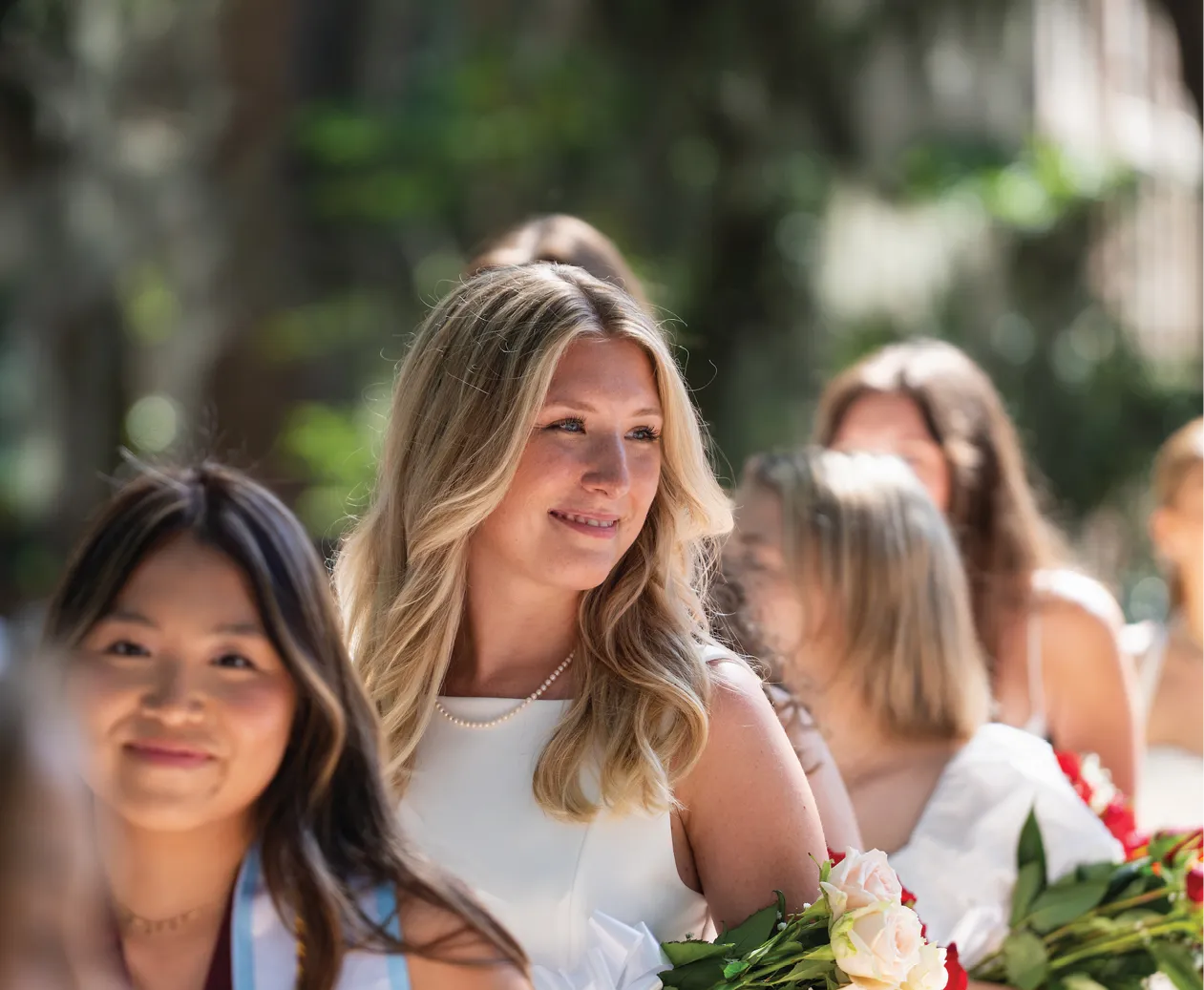A blonde graduate in a sleeveless white dress smiles while walking in a procession. She holds a bouquet of pink and white roses, surrounded by other students in white dresses.