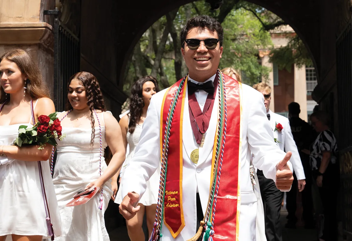 A smiling male graduate in a white tuxedo, black bow tie, and sunglasses gives two thumbs up. He wears colorful cords and a red stole that says “Veteran Peer Advisor” while walking with fellow graduates.