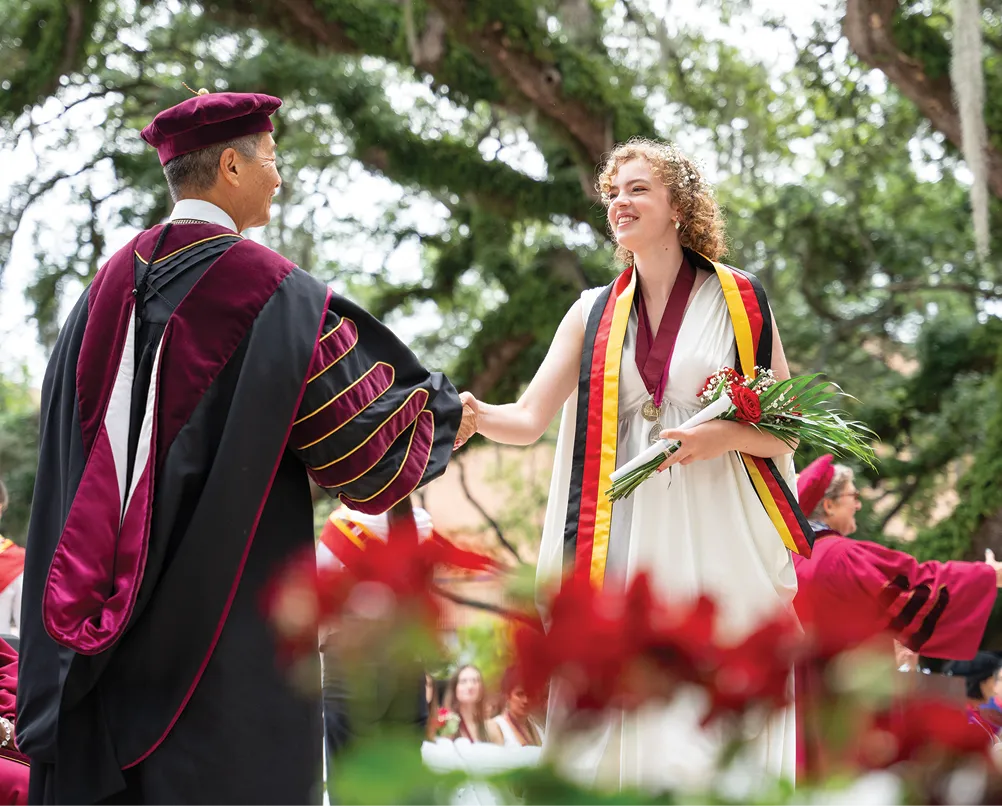 A smiling graduate in a white dress with gold, black, and red stoles shakes hands with a faculty member in maroon and black academic regalia during a graduation ceremony under large oak trees.