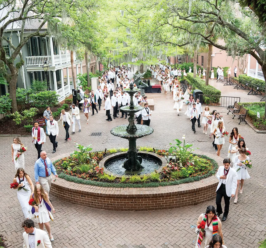 A wide-angle view of graduates in white attire walking through a brick courtyard with a large fountain in the center. The path is lined with trees, plants, and historic campus buildings.