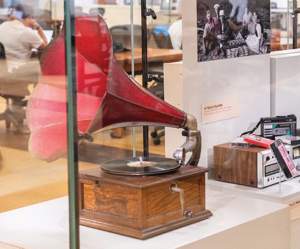 A close-up shot through a glass display case reveals a vintage, dark wood gramophone with a large, bright red horn on the left.