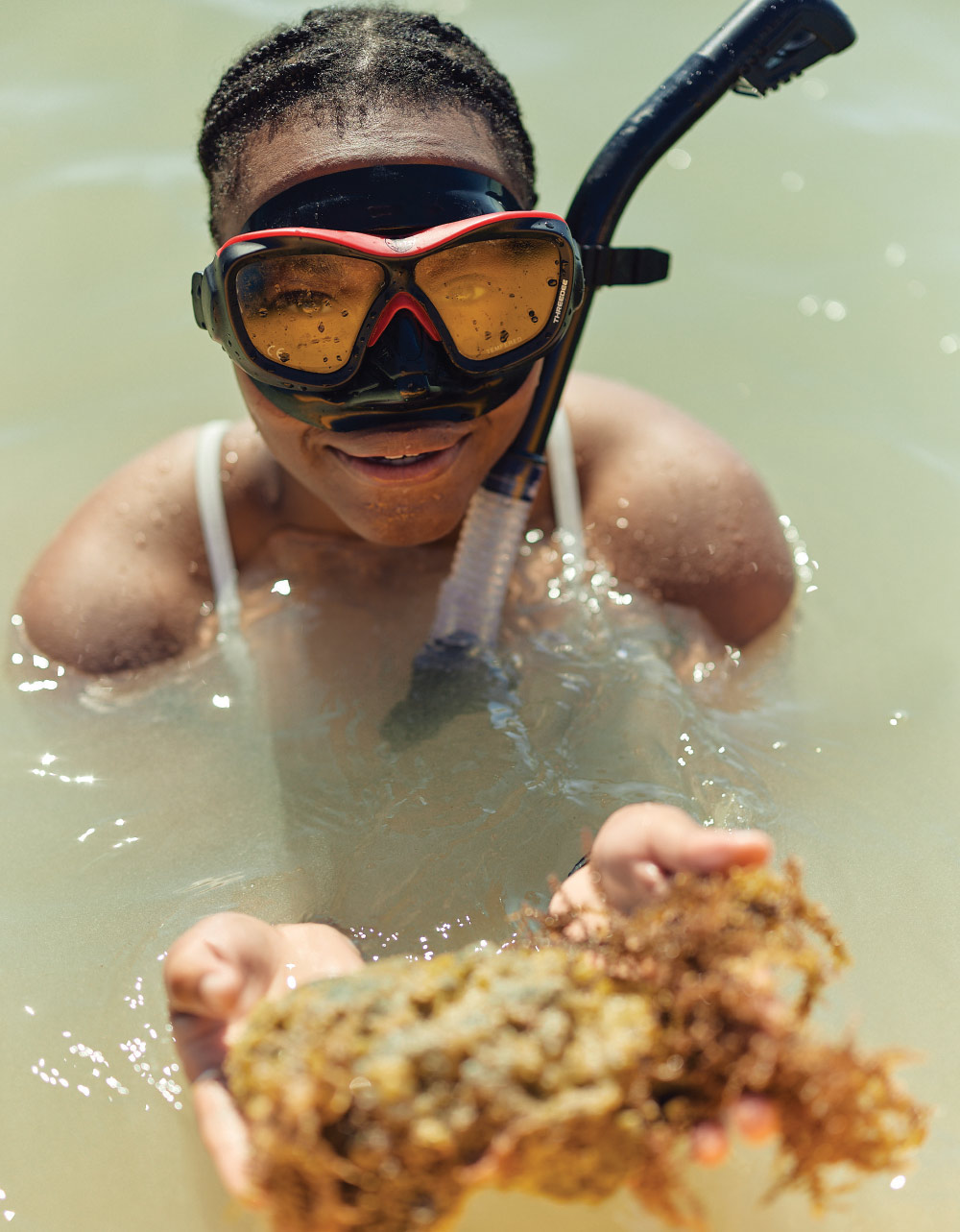Dallas Bryson sitting in shallow water and holding a piece of seaweed while wearing a black and red snorkeling mask with an attached snorkel