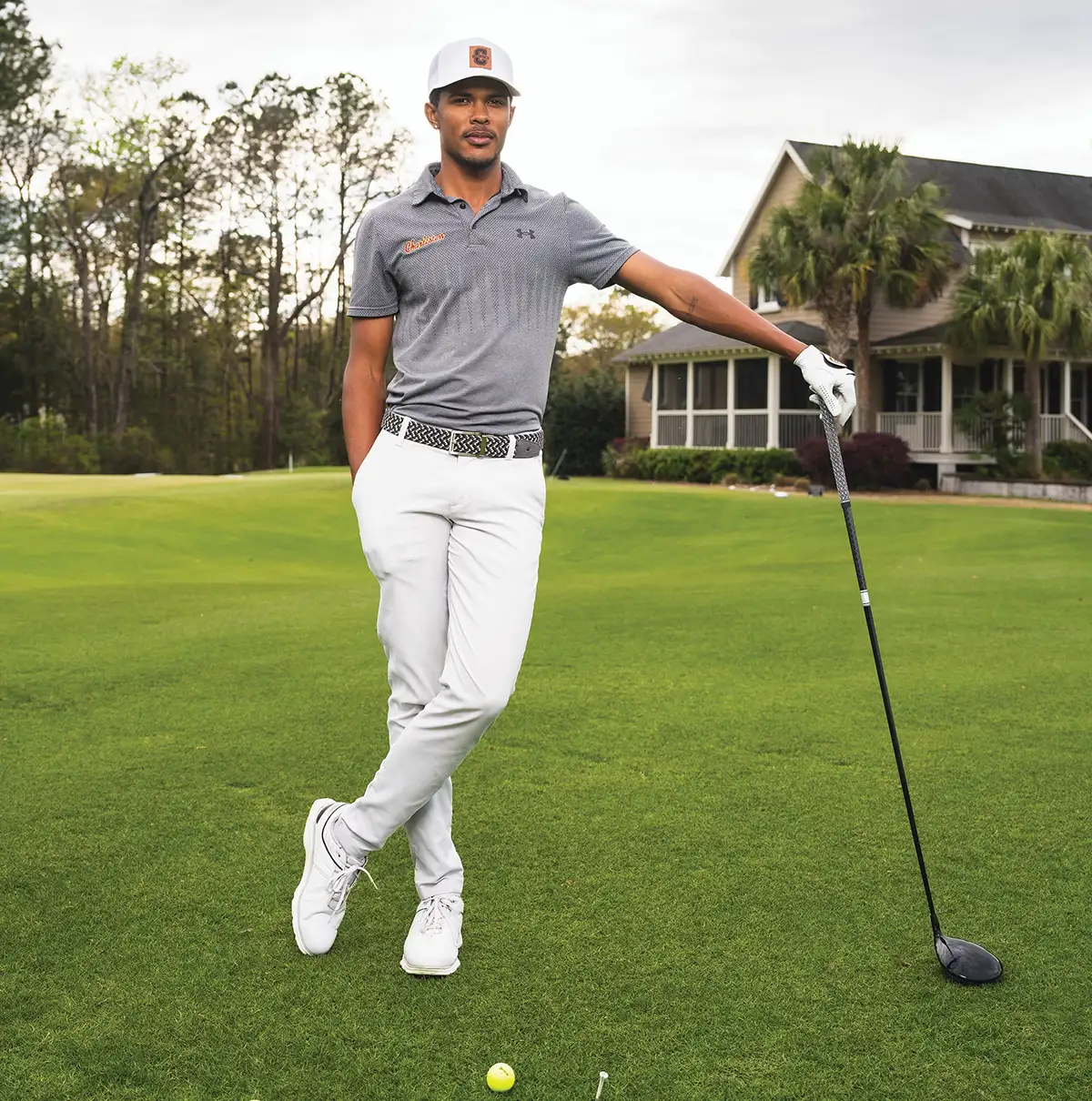 Kieron Van Wyk at a golf course leaning nonchalantly on his golf driver and wearing a white hat, gray polo shirt, white pants, and white shoes