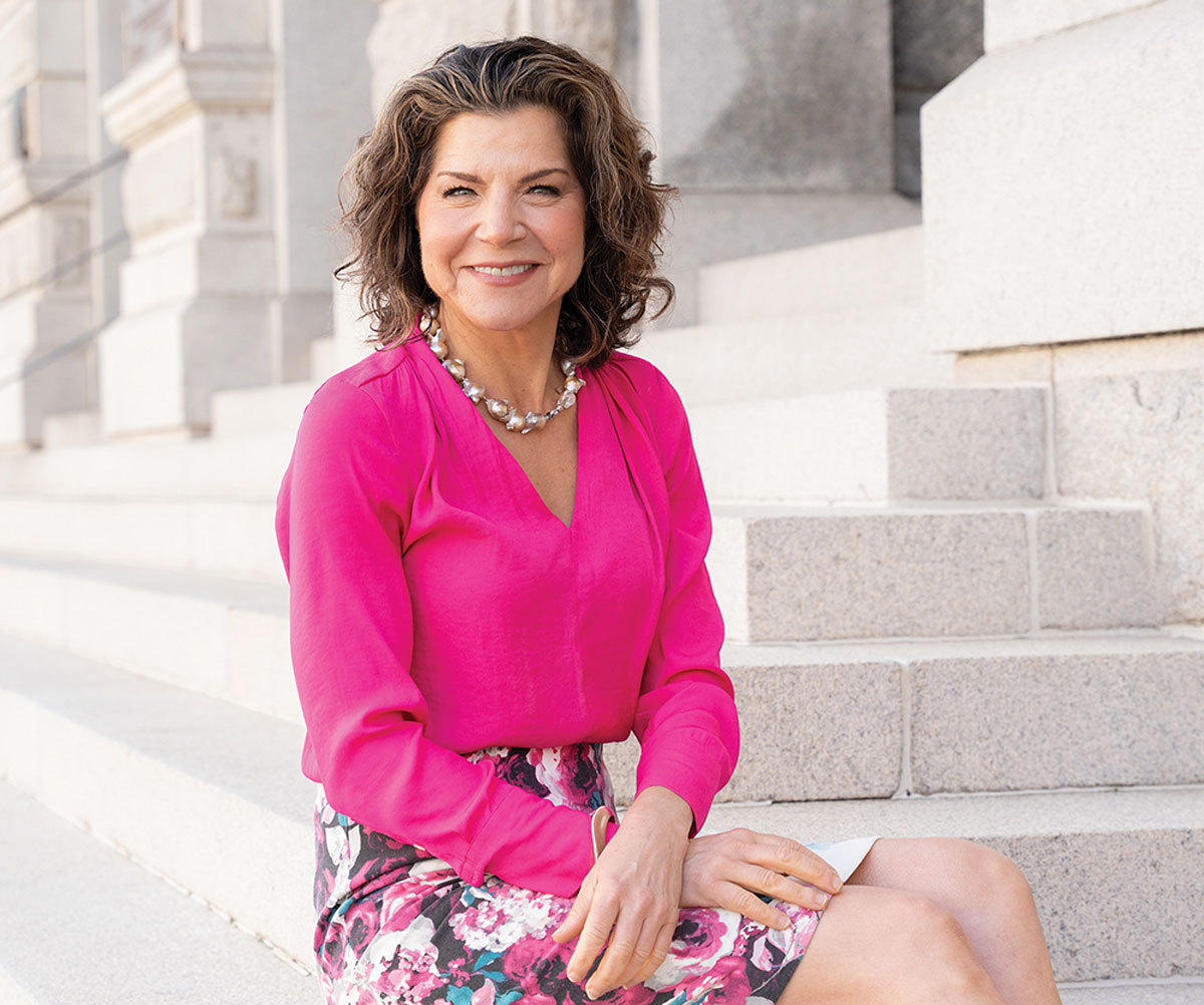 A photo of Claire Wofford smiling while sitting on stone steps. She is wearing a bright pink blouse, a floral skirt, and a pearl-like necklace.