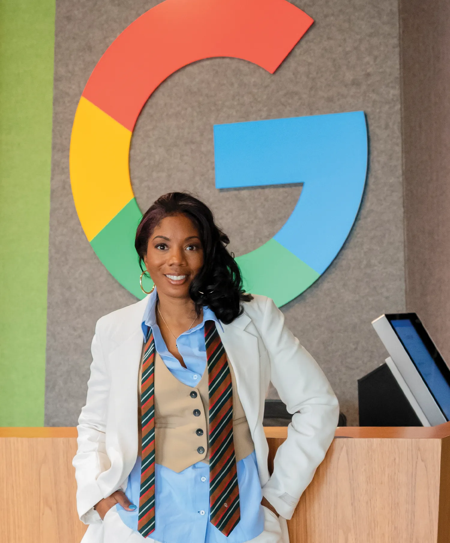 A photo of Rachelle Olden smiling in front of a large, colorful Google "G" logo. She is wearing a white blazer, a light blue shirt, a tan vest, and a striped tie.