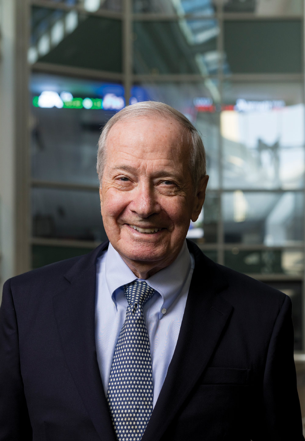 An older man in a suit and tie standing indoors with a friendly expression.