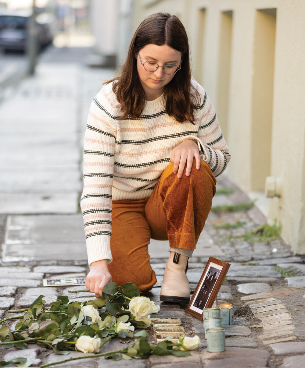 Woman kneeling on a cobblestone street with white roses, a framed photo, and lit candles.