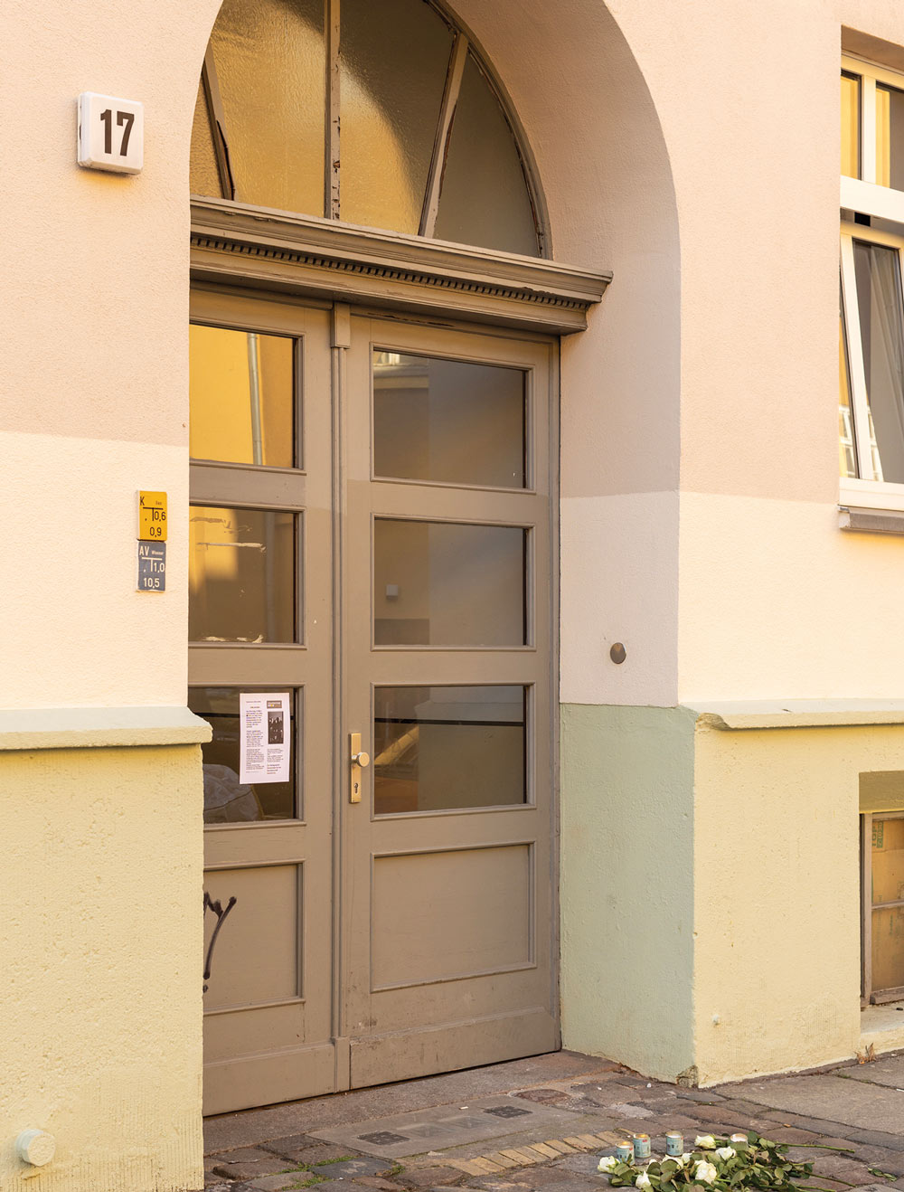 Arched doorway on a beige building with glass-paneled gray door, number 17, a paper on the door, and flowers with candles on the ground.