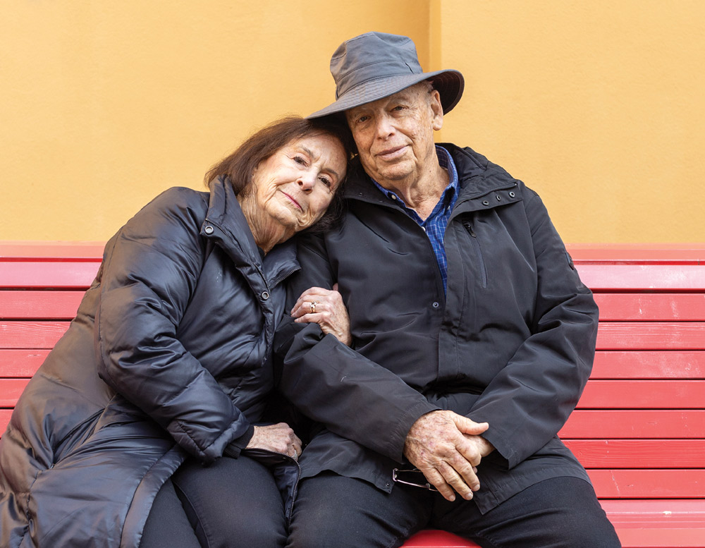 Elderly couple sitting closely on a red bench against a mustard-yellow wall.
