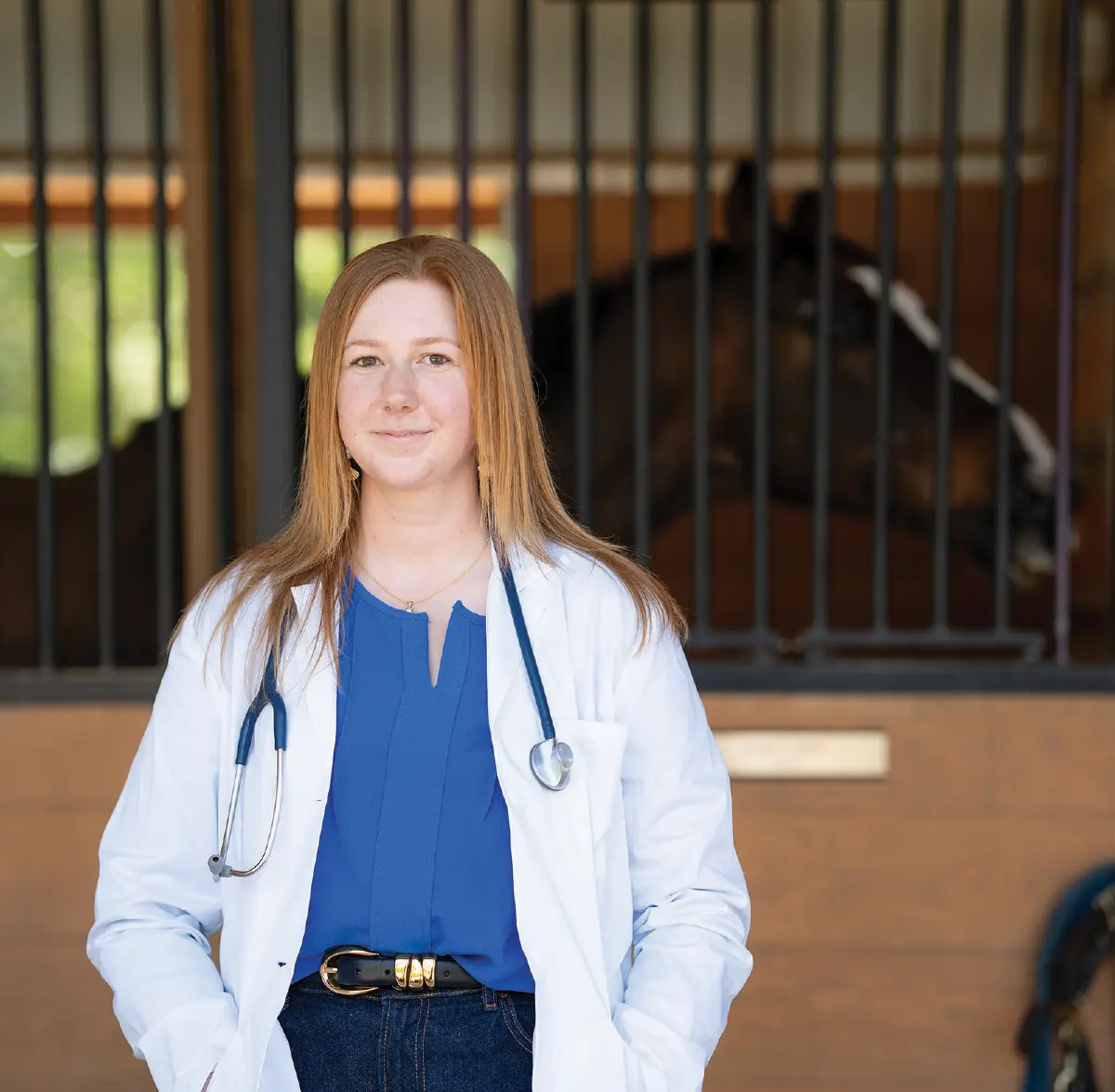 Portrait orientation outdoor photograph of Allyson Pace, a smiling young woman with long reddish-brown hair wearing an open white lab coat over a blue blouse top, dark blue denim jeans, and a stethoscope around her neck as she is standing in a stable with a horse visible in a stall behind her