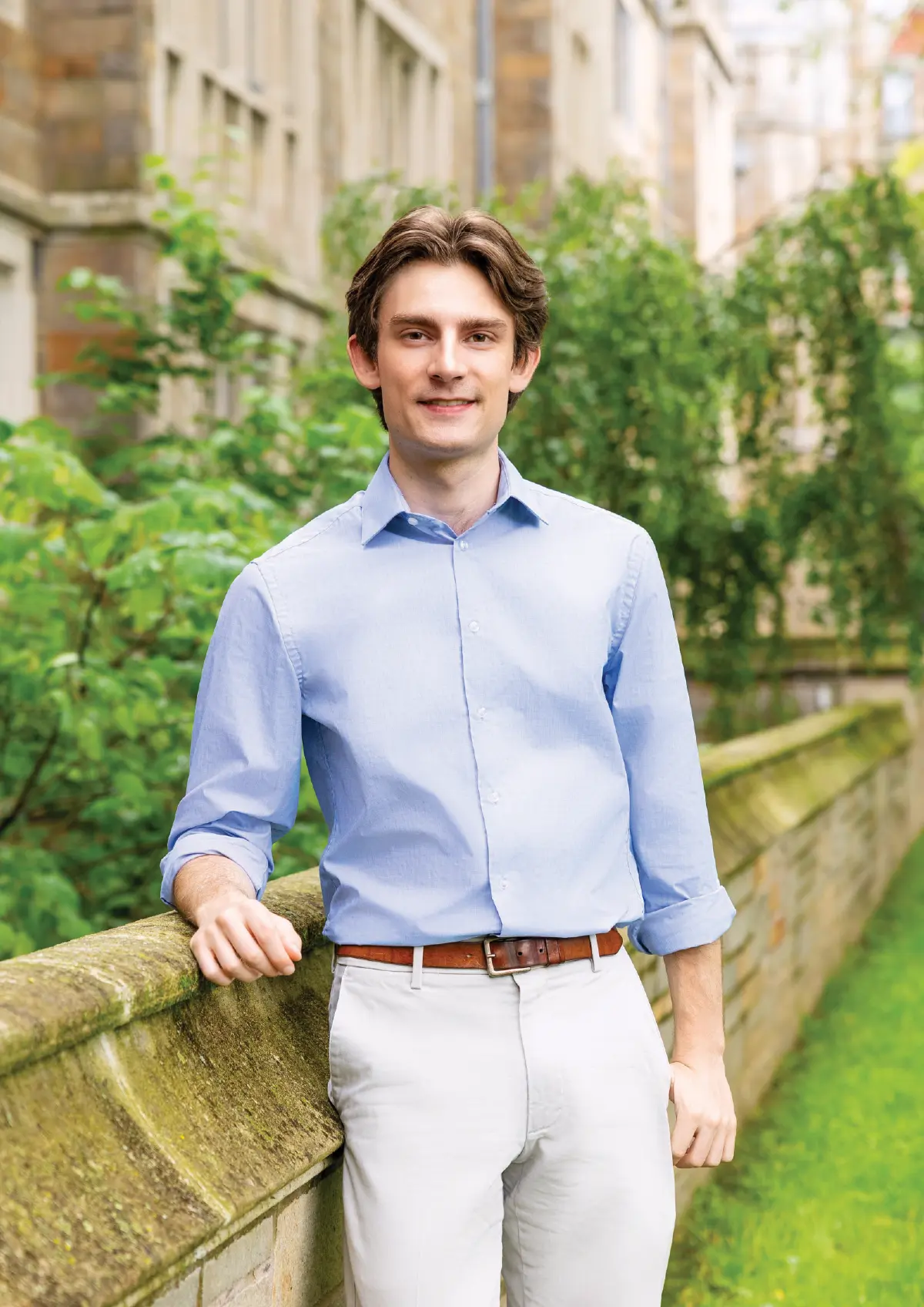 Portrait orientation outdoor photograph of Tom Walter, a smiling young man with dark brown hair, wearing a light sky blue button-down dress shirt with rolled-up sleeves, a brown belt, and light-colored off-white color shade dress pants as he is leaning on a faded mossy rusty stone wall; The blurred background behind him contains green foliage and a stone building
