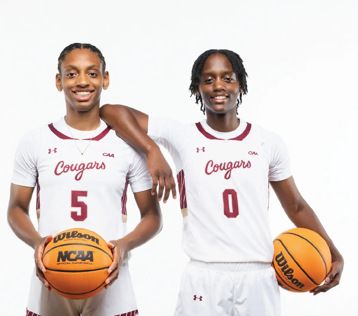 Portrait orientation photograph close-up view of two female basketball players in white Cougars team basketball jerseys with maroon and dark bronze trim outlines in different parts of the jerseys as these individuals stand side-by-side, each holding a Wilson branded basketball containing the NCAA Official Game Ball logo; The player on the left wears number 5 while the player on the right wears number 0. Both are smiling and these women pictured happen to be Taryn and Taylor Barbot