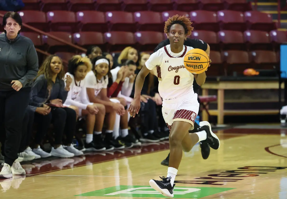 Indoor portrait orientation photograph of Taylor Barbot, who is seen running in a white, dark bronze, and maroon outlined Cougars team jersey with her number 0 visible as she is dribbling a Wilson branded NCAA official game ball logo basketball down the court during a game; Teammates and a coach are visible on the bench in the background around her; Taylor averaged 11.8 points per game