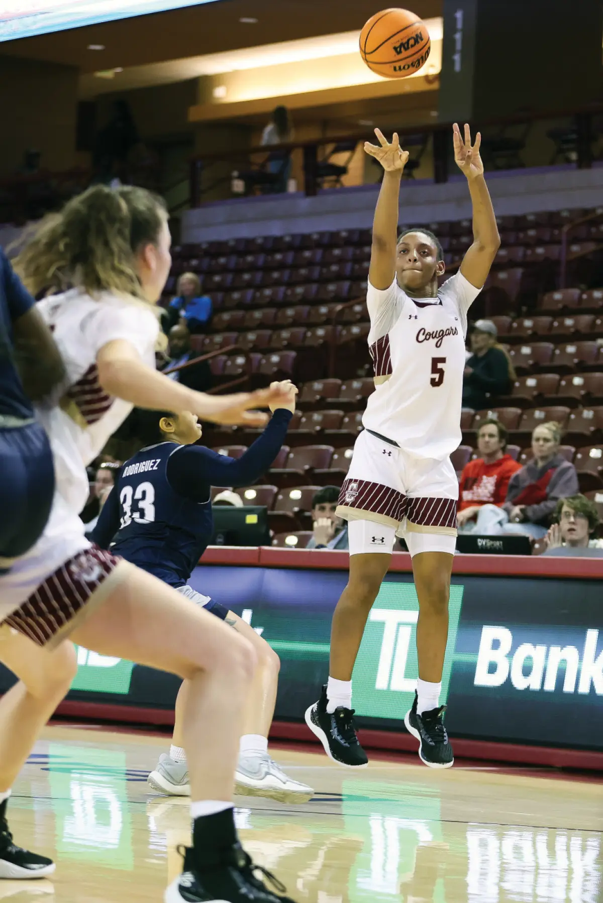 Indoor portrait orientation photograph of Taryn Barbot, who is seen shooting in a white, dark bronze, and maroon outlined Cougars team jersey with her number 5 visible as she is releases a Wilson branded NCAA official game ball logo basketball in motion in air on the court during a game; Other teammates and her opponents are visible on the hardwood floor while there are people seen in the audience gym seats watching the game in the background around her; Taryn is the scoring leader with 17.7 points per game