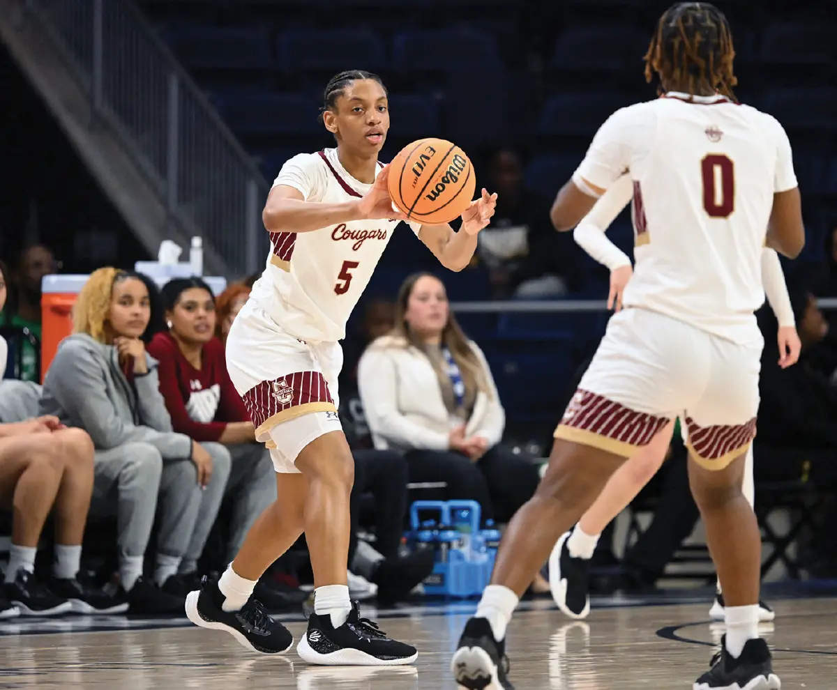 Indoor portrait orientation photograph of Taryn passing to Taylor, who are seen in their respective white, dark bronze, and maroon outlined Cougars team jerseys with their numbers 0 and 5 visibly seen each separately as the Wilson branded NCAA official game ball logo basketball is seen in motion in air on the court during a game; Teammates and a coach are visible on the bench in the background around them