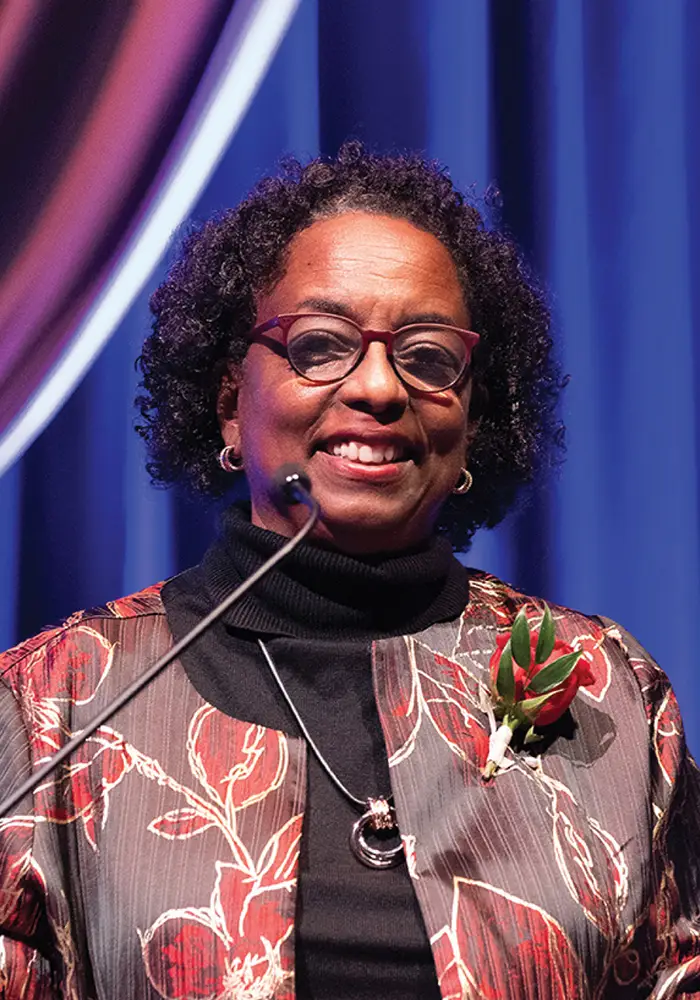 Demetria stands at a podium on a stage, smiling while speaking into a microphone, wearing glasses and a patterned jacket with a red corsage, with blue stage drapery behind her.