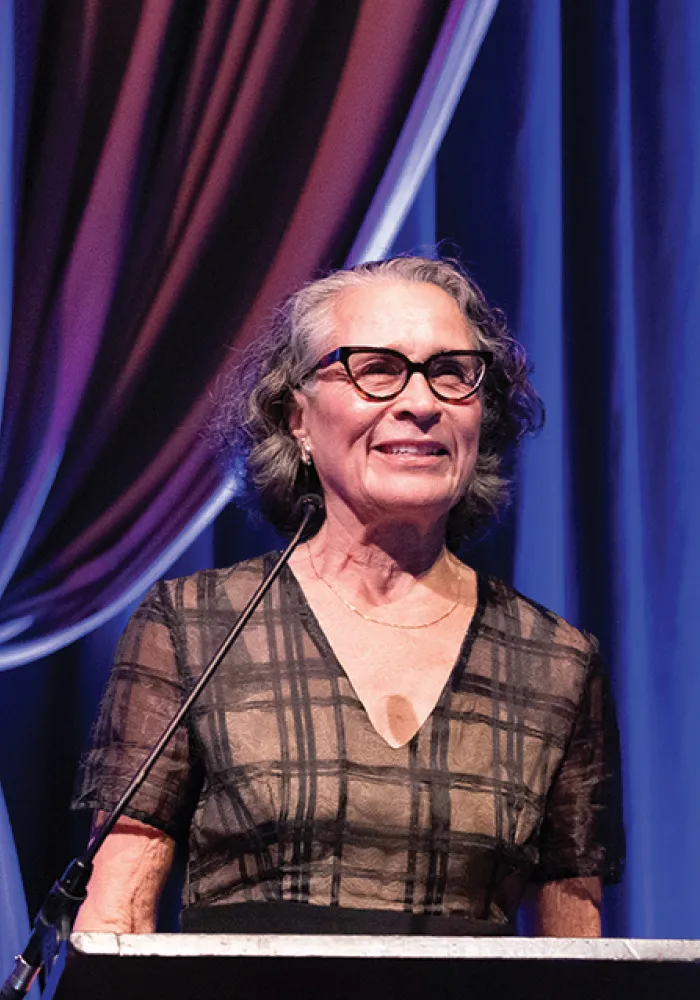 Arlinda smiles while speaking at a podium onstage, wearing glasses and a dark patterned dress, with softly lit blue and purple curtains in the background.