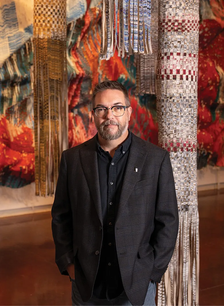 A man wearing glasses and a dark blazer stands in an art gallery in front of large, colorful woven textile installations that hang from the ceiling and walls.