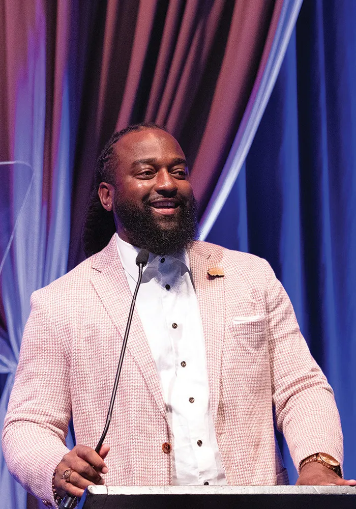 Marcus stands at a podium addressing an audience, wearing a light-colored blazer over a white shirt, smiling as he speaks under blue stage lighting.
