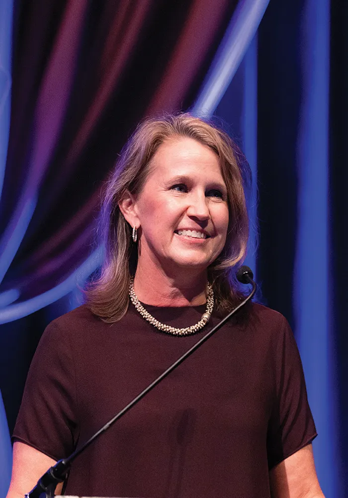 Cynthia speaks at a podium onstage, wearing a maroon dress and pearl necklace, smiling slightly as she looks toward the audience against blue draped curtains.