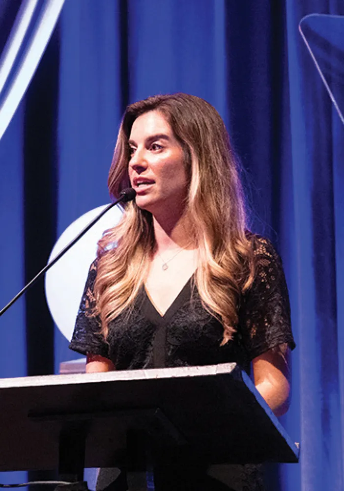 Deniz speaks at a podium onstage, wearing a black lace dress and looking to the side as she addresses the audience, framed by blue curtains and stage lighting.