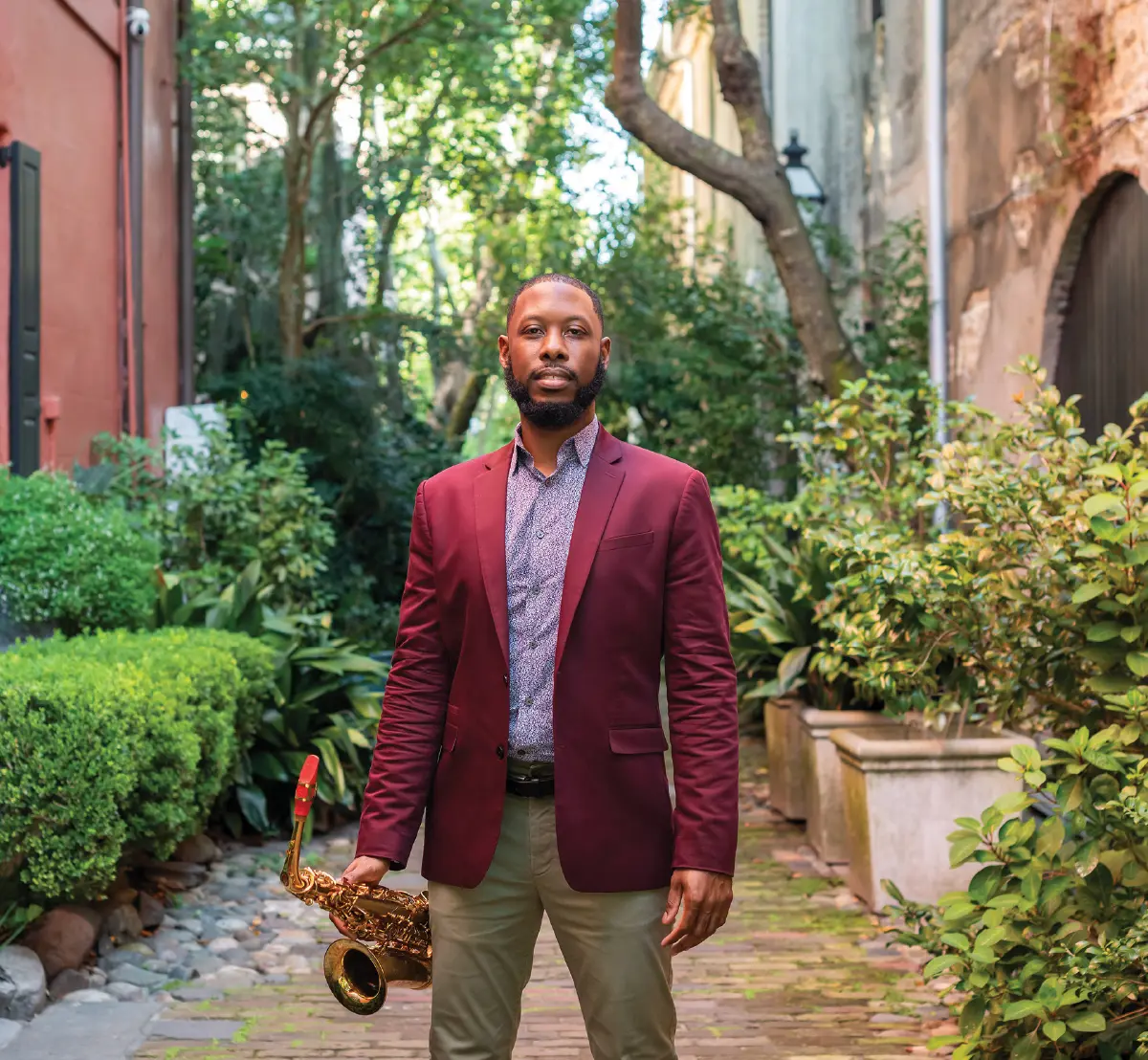 Kevin Patton, a smiling man with a beard wearing a maroon blazer and a patterned shirt, stands in a narrow, lush alleyway paved with bricks; He is holding a gold saxophone in his right hand