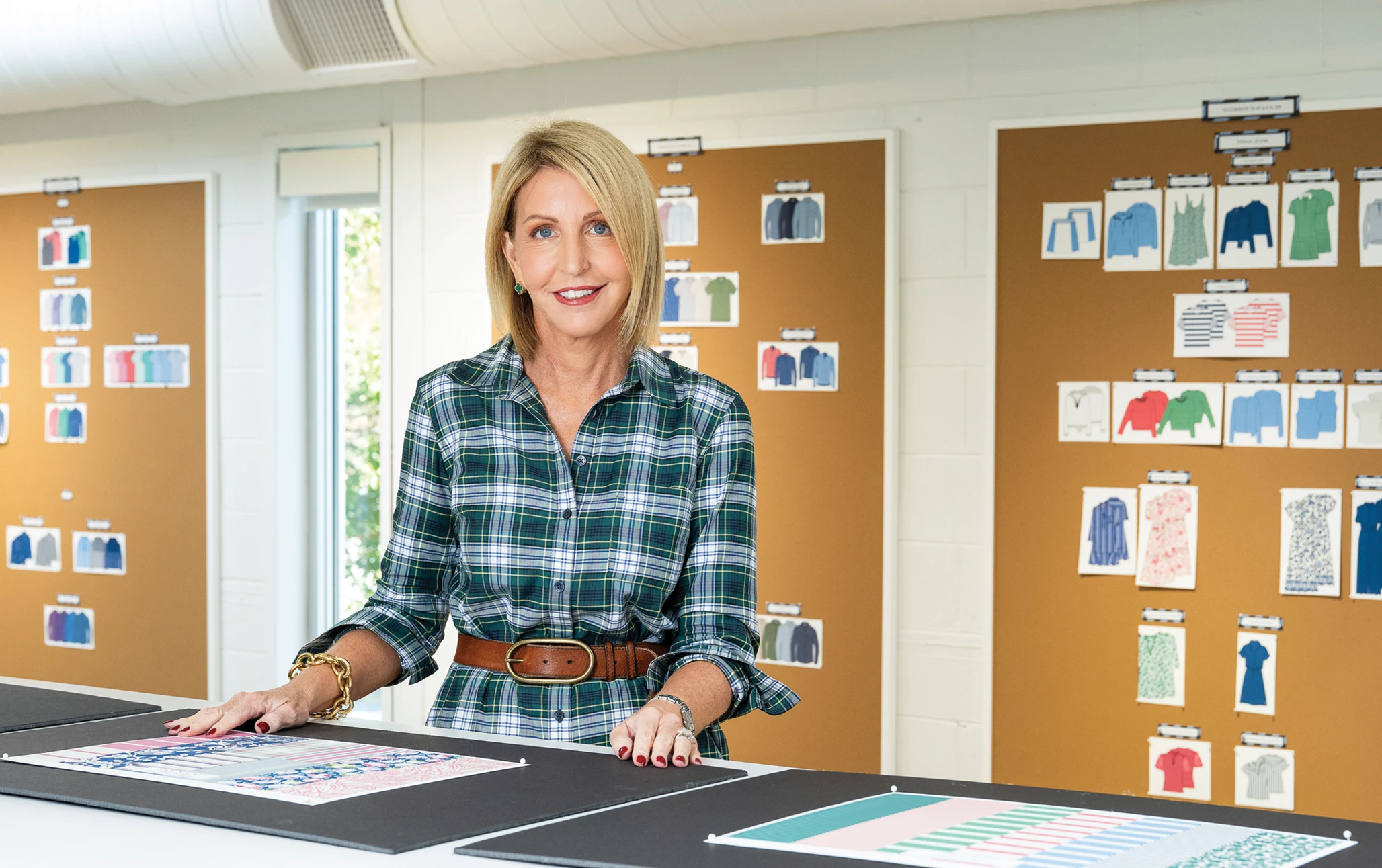 A woman wearing a green plaid dress stands at a design worktable in a studio, smiling at the camera, with fabric samples and color layouts on the table and clothing sketches pinned to large cork boards behind her.