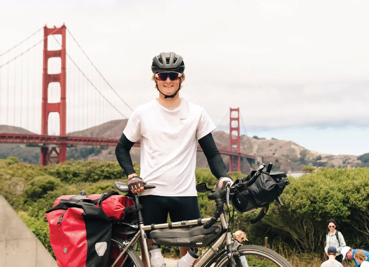 Ben Navarro, a smiling male cyclist guy wearing a helmet, sunglasses, and a white t-shirt, stands with a loaded touring bike; The Golden Gate Bridge is prominently visible in the background under a bright, overcast sky with a few other random people standing nearby closer to some greenery bushes behind Ben Navarro