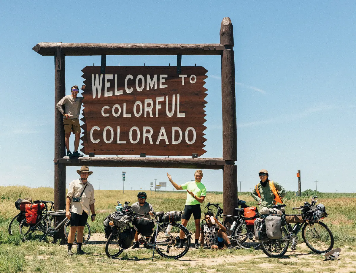 Six male cyclists pose for a photo in front of a large wooden Welcome to Colorful Colorado sign; They are standing with their touring bikes in a flat, grassy landscape under a clear sunny blue sky